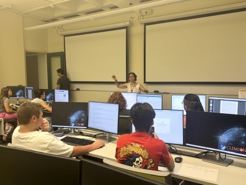 A woman stands at the front of a classroom talking to a group of students, who are all sitting at computers.