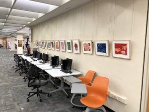 14 paintings hang on a white wall above a desk of computers