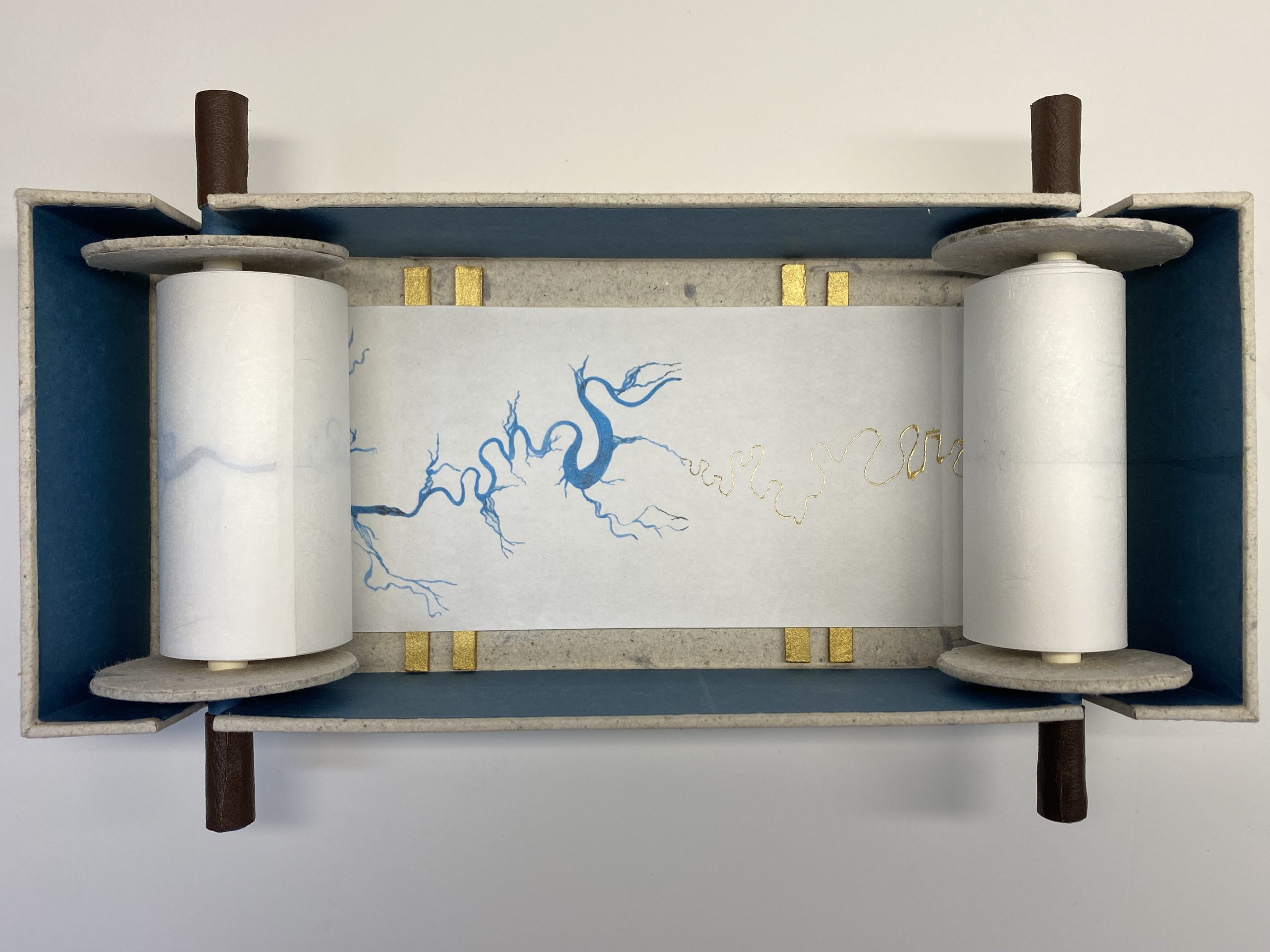 A white paper scroll that has been unfurled and is set in a rectangular blue box. On the scroll there is an abstract drawing done in blue ink and gold foiling. 